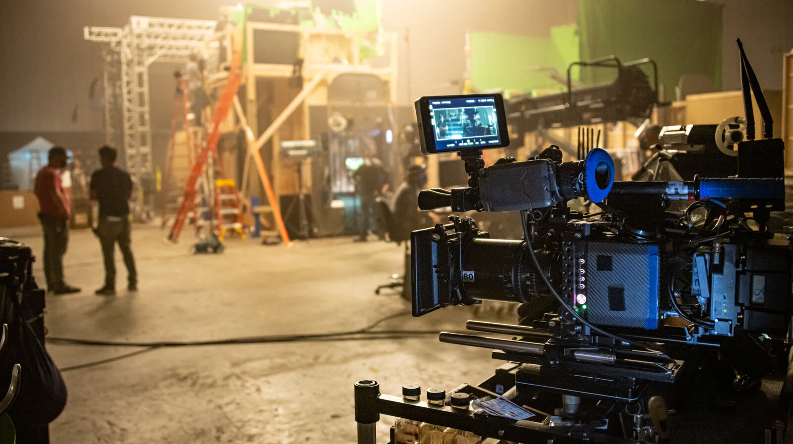 A cinema camera rig on a production set, with crew at work among scaffolding and a green screen behind