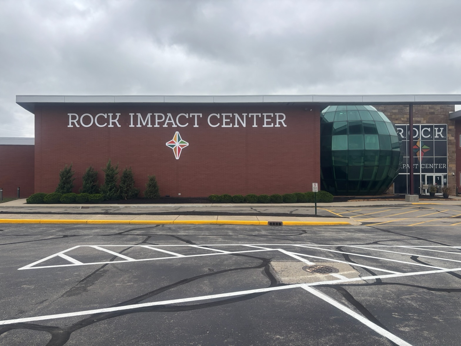 The ROCK Impact Center's brick façade on an overcast day — ROCK IMPACT CENTER in white lettering over the four-point star emblem, a row of young evergreens below, the Globe Room's green glass dome at the corner, and the stone entry tower beyond the parking lot